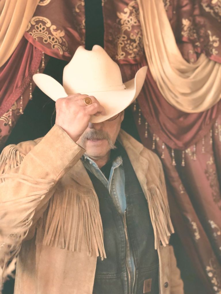 George Ray Russell in a tasseled beige suede coat holding an ecru cowboy hat down over his eyes. He stands before a sumptuous curtain.